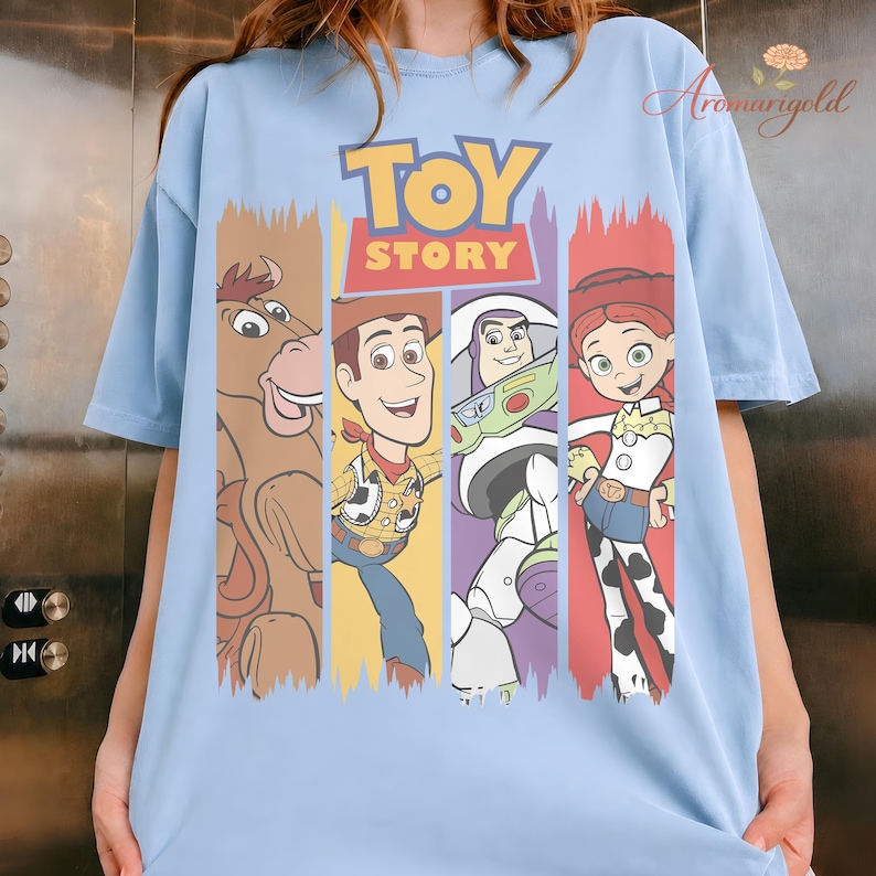 Puede incluir: Camiseta azul claro con las palabras "TOY STORY" en amarillo y rojo, y personajes de dibujos animados. El dise&ntilde;o incluye un caballo marr&oacute;n, Woody, Buzz Lightyear y Jessie, cada uno en un bloque de color vertical.