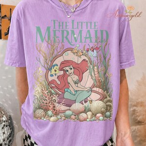 May include: A lilac-colored t-shirt featuring a graphic of Ariel from "The Little Mermaid." The design includes Ariel in a clam shell, with Flounder, Sebastian, and other sea creatures. The words "The Little Mermaid" are printed above the image.