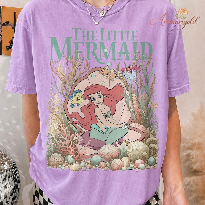 May include: A lilac-colored t-shirt featuring a graphic of Ariel from "The Little Mermaid." The design includes Ariel in a clam shell, with Flounder, Sebastian, and other sea creatures. The words "The Little Mermaid" are printed above the image.
