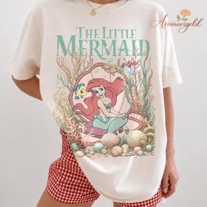 May include: Cream-colored t-shirt featuring a colorful graphic of Ariel from The Little Mermaid. The design includes Ariel, Flounder, and Sebastian, along with the words "The Little Mermaid" in a teal font. The shirt is a casual, comfortable style.