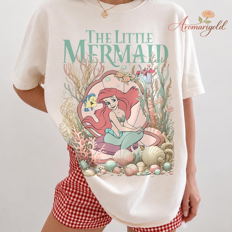 May include: Cream-colored t-shirt featuring a colorful graphic of Ariel from The Little Mermaid. The design includes Ariel, Flounder, and Sebastian, along with the words "The Little Mermaid" in a teal font. The shirt is a casual, comfortable style.