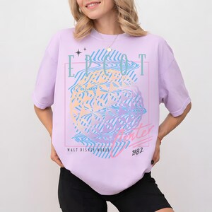 May include: A light purple t-shirt with a graphic design featuring the text "EPCOT Center" and "1982" in a retro style. The design includes a geometric sphere with wavy lines in shades of blue, yellow, and pink. The words "Walt Disney World" are also present.
