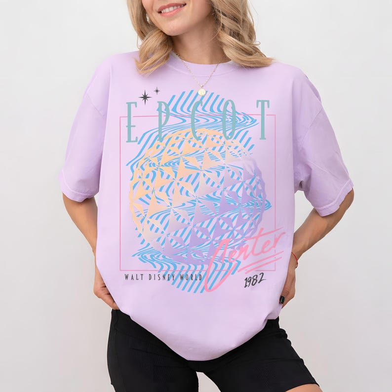 May include: A light purple t-shirt with a graphic design featuring the text "EPCOT Center" and "1982" in a retro style. The design includes a geometric sphere with wavy lines in shades of blue, yellow, and pink. The words "Walt Disney World" are also present.