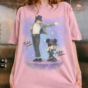 May include: Pink t-shirt featuring a graphic of Michael Jackson and Mickey Mouse in matching outfits. The design includes the text "Michael Jackson" and "Mickey Mouse" in cursive, set against a purple and blue background with stars.