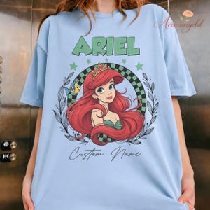 May include: Pale blue t-shirt with a graphic of Ariel from The Little Mermaid. The design includes the name "ARIEL" in green, a portrait of Ariel, and the words "Custom Name". The graphic is surrounded by a checkered pattern and stars.