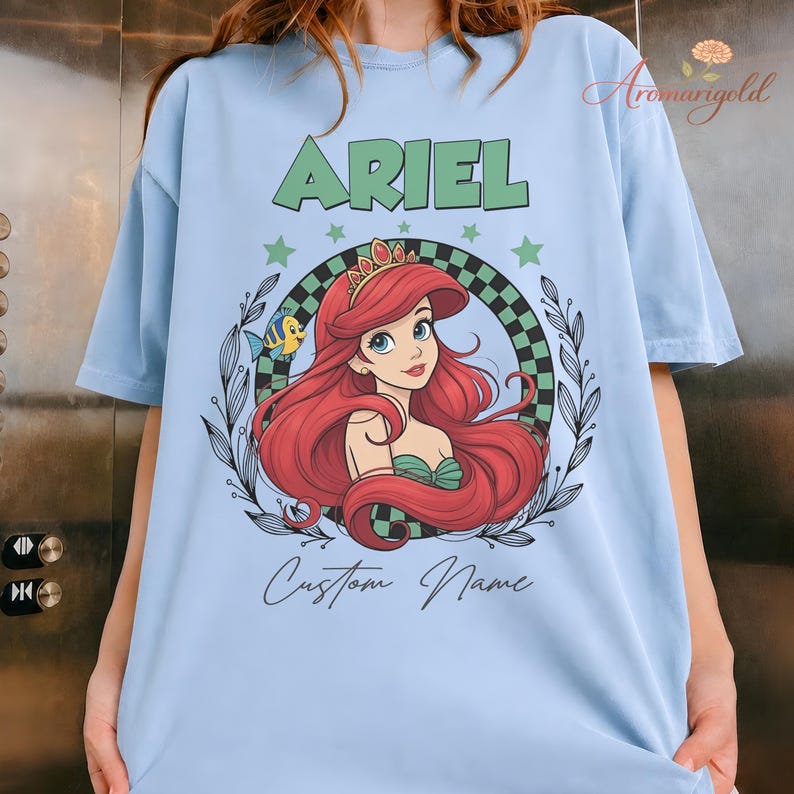 May include: Pale blue t-shirt with a graphic of Ariel from The Little Mermaid. The design includes the name "ARIEL" in green, a portrait of Ariel, and the words "Custom Name". The graphic is surrounded by a checkered pattern and stars.