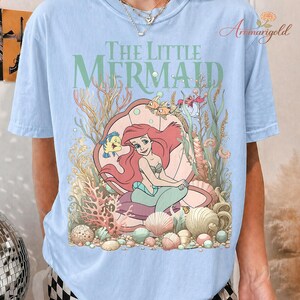 May include: Light blue t-shirt featuring a colorful graphic of Ariel from The Little Mermaid. The design includes Ariel, Flounder, and other sea creatures, with the words "The Little Mermaid" in green.