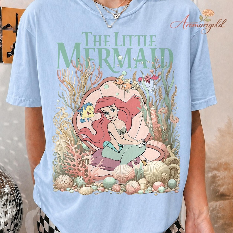 May include: Light blue t-shirt featuring a colorful graphic of Ariel from The Little Mermaid. The design includes Ariel, Flounder, and other sea creatures, with the words "The Little Mermaid" in green.