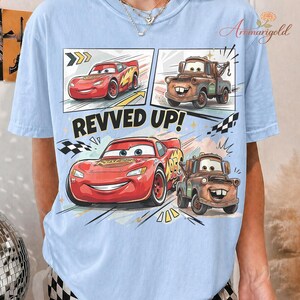 May include: Light blue t-shirt featuring a graphic of Lightning McQueen and Mater from the Cars movie. The design includes comic book style panels with the text "REVVED UP!" in bold letters. The shirt is made of a soft material.