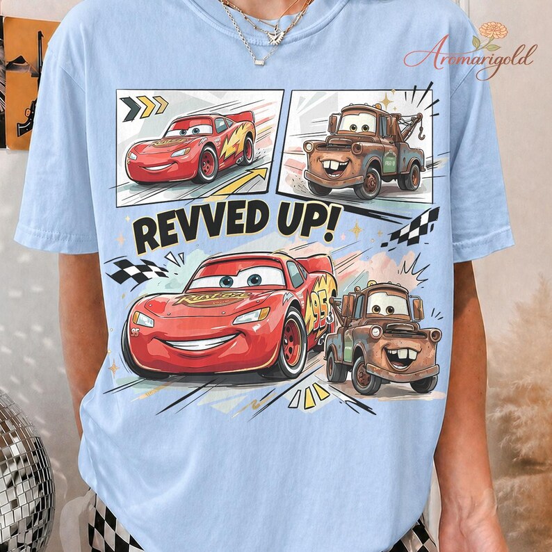May include: Light blue t-shirt featuring a graphic of Lightning McQueen and Mater from the Cars movie. The design includes comic book style panels with the text "REVVED UP!" in bold letters. The shirt is made of a soft material.