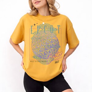May include: Mustard yellow t-shirt featuring a graphic design with the text "EPCOT Center" in a retro style. The design includes a stylized globe and the text "Walt Disney World 1982".