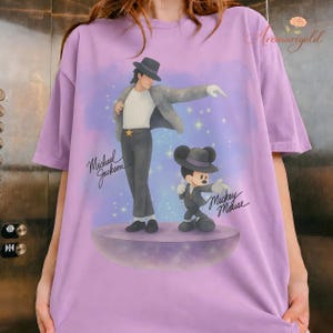 May include: A light purple t-shirt featuring a graphic of Michael Jackson and Mickey Mouse in matching outfits. The text "Michael Jackson" and "Mickey Mouse" are written in cursive. The background is a purple and blue gradient with stars.