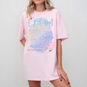 May include: A light pink t-shirt with a graphic design featuring the text "EPCOT Center" and "Winter 1982" in a retro style. The design includes a colorful, wavy globe-like image. The shirt is a casual, oversized fit.