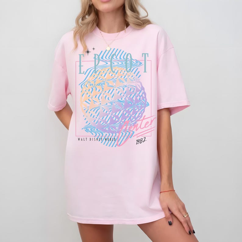 May include: A light pink t-shirt with a graphic design featuring the text "EPCOT Center" and "Winter 1982" in a retro style. The design includes a colorful, wavy globe-like image. The shirt is a casual, oversized fit.