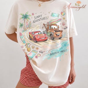 以下が含まれることがあります： クリーム色のTシャツ。映画「カーズ」のライトニング・マックィーンとメーターのカラフルな漫画イラストがビーチに描かれています。「SUNNY ADVENTURES!」と「BEACHSIDE MATES!」の文字も含まれています。