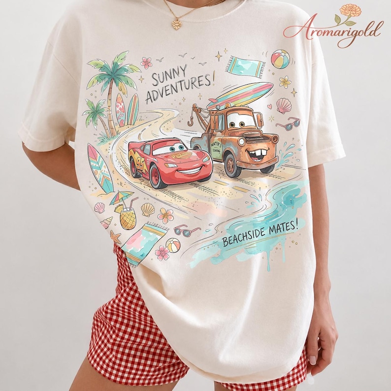 以下が含まれることがあります： クリーム色のTシャツ。映画「カーズ」のライトニング・マックィーンとメーターのカラフルな漫画イラストがビーチに描かれています。「SUNNY ADVENTURES!」と「BEACHSIDE MATES!」の文字も含まれています。