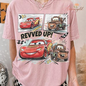 May include: Pink t-shirt featuring a comic strip design with the characters Lightning McQueen and Mater from the movie Cars. The text "REVVED UP!" is displayed prominently. The shirt has a crew neck and short sleeves.
