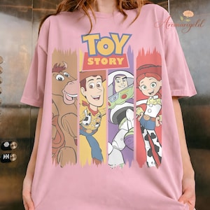 Puede incluir: Camiseta rosa claro con las palabras "TOY STORY" en amarillo y rojo, con ilustraciones de dibujos animados de Woody, Buzz Lightyear, Jessie y Bullseye. La camiseta tiene mangas cortas y cuello redondo.