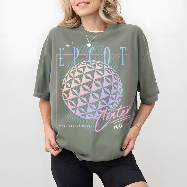 May include: Olive green t-shirt featuring the text "EPCOT Center" with a geometric sphere design in pastel colors. The shirt also includes "Walt Disney World" and the year "1982".