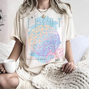 May include: Cream-colored t-shirt featuring a colorful graphic design with the text "EPCOT Center" and "Walt Disney World 1982". The graphic includes a geometric sphere in shades of blue, purple, and orange.