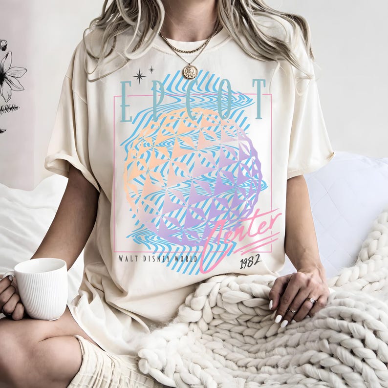 May include: Cream-colored t-shirt featuring a colorful graphic design with the text "EPCOT Center" and "Walt Disney World 1982". The graphic includes a geometric sphere in shades of blue, purple, and orange.