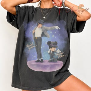 May include: Dark gray t-shirt featuring a graphic of Michael Jackson and Mickey Mouse in matching outfits. The design includes the text "Michael Jackson" and "Mickey Mouse" in a stylized font, set against a purple and blue background.