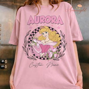 May include: Pale pink t-shirt with a graphic of Princess Aurora, the word "AURORA", and the text "Custom Name". The design includes a floral wreath and stars.