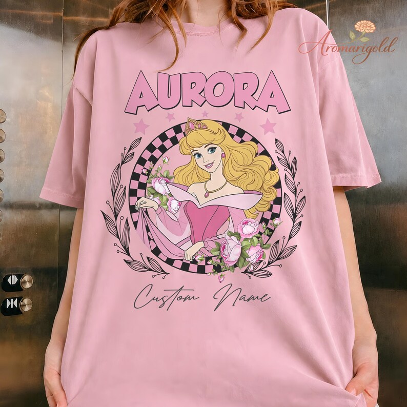 May include: Pale pink t-shirt with a graphic of Princess Aurora, the word "AURORA", and the text "Custom Name". The design includes a floral wreath and stars.