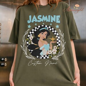 May include: An olive green t-shirt with a graphic of Princess Jasmine. The name "JASMINE" is in light blue above her. The design includes a checkered circle, stars, and the words "Custom Name" below the image.