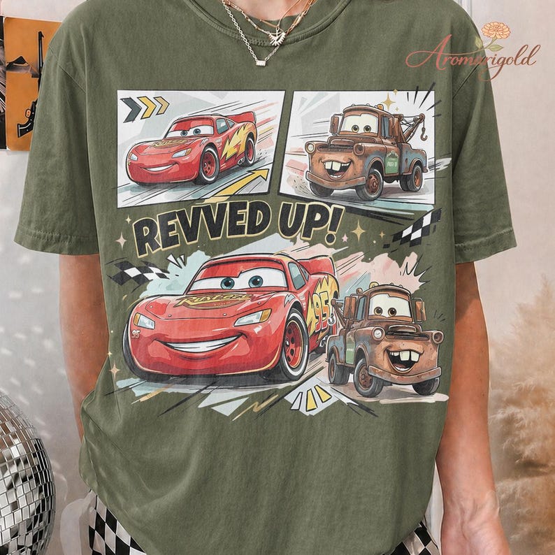 May include: Olive green t-shirt featuring a graphic of Lightning McQueen and Mater from the movie Cars. The design includes the text "REVVED UP!" and racing-themed elements. The shirt is a casual wear item.