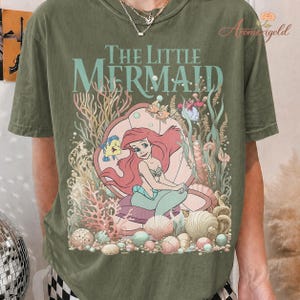 May include: Olive green t-shirt featuring a graphic of Ariel from The Little Mermaid. The design includes the text "The Little Mermaid" in a seafoam green color, along with illustrations of sea creatures and coral.