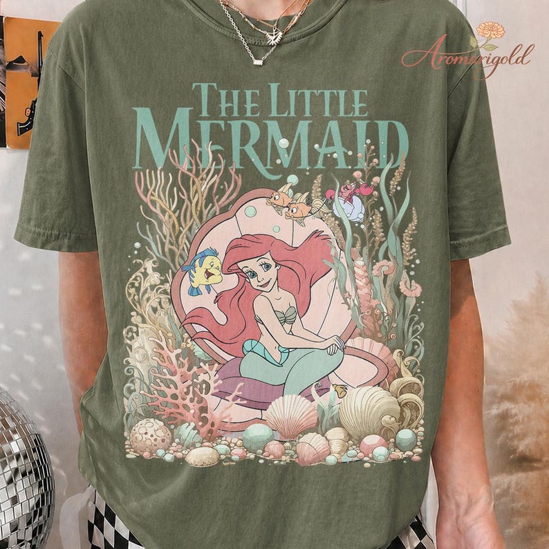 May include: Olive green t-shirt featuring a graphic of Ariel from The Little Mermaid. The design includes the text "The Little Mermaid" in a seafoam green color, along with illustrations of sea creatures and coral.
