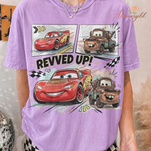 May include: A light purple t-shirt featuring a comic strip design with the text "REVVED UP!". The design includes illustrations of the Cars characters Lightning McQueen and Mater.