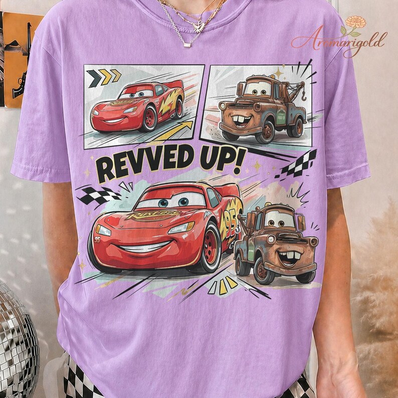 May include: A light purple t-shirt featuring a comic strip design with the text "REVVED UP!". The design includes illustrations of the Cars characters Lightning McQueen and Mater.