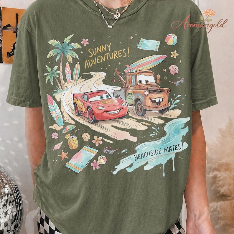 以下が含まれることがあります： オリーブグリーンのTシャツ。映画「カーズ」のライトニング・マックィーンとメーターのグラフィックが特徴で、「Sunny Adventures!」と「Beachside Mates!」の文字が入っています。ヤシの木、サーフボード、ビーチの要素が含まれています。