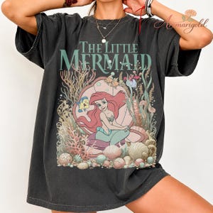 May include: Dark gray t-shirt featuring a graphic of Ariel from The Little Mermaid. The design includes Ariel, Flounder, and various sea elements. The text "The Little Mermaid" is printed above the illustration in a teal color.