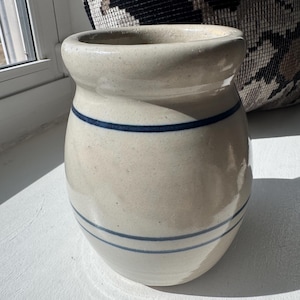 May include: A cream-colored ceramic jar with two navy blue stripes. The jar has a rounded shape and a slightly flared rim. The jar is sitting on a white surface.
