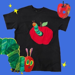May include: Black t-shirt featuring a colorful applique of a red apple with a green worm and a green leaf. The shirt also has a partial illustration of a green and yellow caterpillar and a red apple. The background is blue.