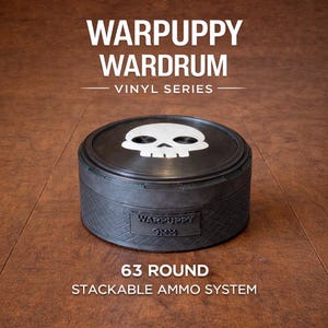 May include: A black WARPUPPY WARDRUM vinyl series ammo container with a white skull design on top. The container is labeled "9MM" and "63 ROUND STACKABLE AMMO SYSTEM". The container is cylindrical and sits on a wooden surface.