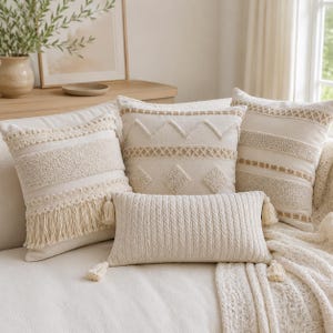 May include: A collection of four cream-colored throw pillows. Three square pillows feature textured patterns including fringe, zigzags, and woven stripes. One rectangular lumbar pillow has a vertical cable knit design with tassels on each corner.
