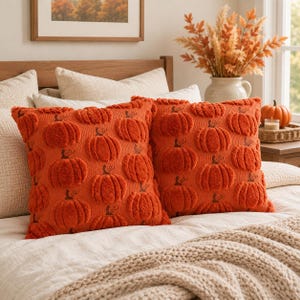 May include: Two square throw pillows in a vibrant orange color, featuring a repeating pattern of raised, textured pumpkins. The pumpkins are knitted with a fuzzy, plush surface and include small embroidered stems and vines on each individual motif.