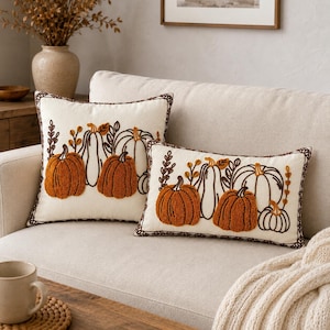 Puede incluir: Dos cojines decorativos de color crema con diseños de calabazas bordadas en relieve en tonos naranja y marrón. Tanto el cojín cuadrado como el rectangular muestran motivos detallados de calabazas con tallos y bordes marrón oscuro.