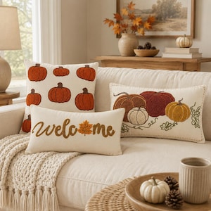 May include: Three decorative throw pillows with cream-colored fabric and textured embroidery. One square pillow features orange pumpkins, one rectangular pillow displays the word welcome with a leaf, and one rectangular pillow shows pumpkins.