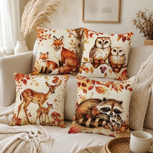 May include: A set of four square throw pillows featuring woodland animal illustrations. One pillow shows a fox and kit, another two owls on a branch, one a deer and fawn, and the last a raccoon and kit, all surrounded by autumn leaves and foliage.