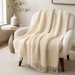 Puede incluir: Una manta de color crema con un patrón texturizado de rayas verticales acanaladas alternadas y cuadrados de relieve, terminada con flecos largos en el borde inferior, colocada sobre un sillón tapizado de color blanco roto.