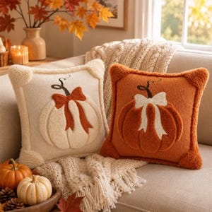 Puede incluir: Dos cojines cuadrados con diseños de calabaza de felpa en relieve. Uno es de color crema con un lazo naranja óxido, mientras que el otro es naranja óxido con un lazo crema. Ambos cojines tienen una tela texturizada y suave al tacto.