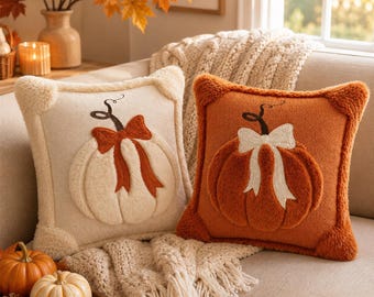 May include: Two square throw pillows featuring plush, raised pumpkin designs. One pillow is cream-colored with a rust-orange bow, while the other is rust-orange with a cream-colored bow. Both pillows have textured, fuzzy fabric and corner accents.