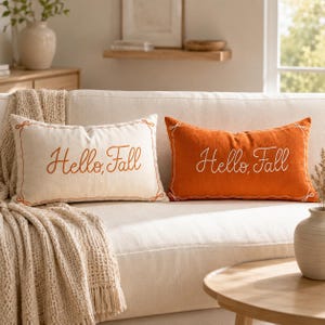 Op de afbeelding: Twee rechthoekige sierkussens met de tekst Hello, Fall. Het ene kussen is crème met oranje tekst en een oranje rand, het andere is oranje met witte tekst en een witte rand. Beide kussens hebben een eenvoudig lijnontwerp als kader.