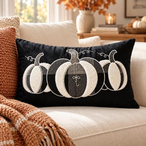 May include: A rectangular black throw pillow featuring an embroidered design of three pumpkins. The pumpkins are rendered in white and black with textured, contrasting patterns including dots, checks, and solid sections, with curved stems.