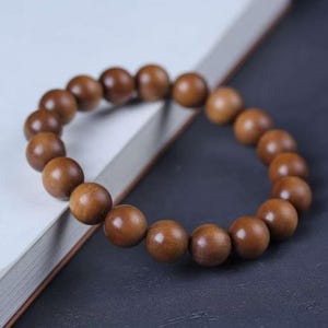 Könnte beinhalten: Ein kreisförmiges Armband aus glatten, polierten, mittelbraunen Holzperlen, die aneinandergereiht sind. Die Perlen sind in Größe und Form einheitlich und weisen eine natürliche Holzmaserung mit subtilen Farbvariationen auf jeder einzelnen Perle auf.
